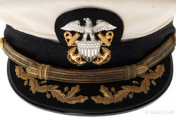 A U.S. Navy officer’s white combination cap with a black visor embroidered with gold oak leaves and acorns. The cap features a silver eagle insignia with crossed anchors and a gold chin strap held by buttons engraved with the Navy emblem.