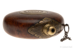 A circular powder flask from the late 18th century, possibly Russian or Eastern European. The burled walnut body features a central brass medallion with engraved patterns, a brass pouring spout with leaf-like engravings, and a bone or ivory base plug